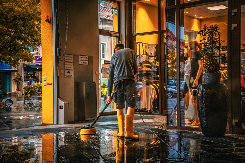 A person wearing yellow rubber boots and casual clothing is engaged in surface cleaning outside a retail store in Woking town centre, Surrey, using a pressure washer to remove dirt and grime from the